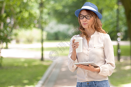 快乐的轻女人着次咖啡杯,手里着平板电脑,喝着咖啡,着夏天的公园背景微笑图片