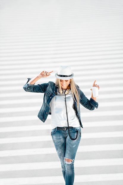 漂亮的轻女人,戴着帽子牛仔裤夹克,带着外卖咖啡杯,站马路上骑着斑马