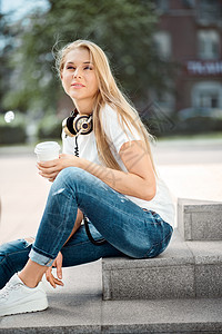 快乐的轻女人戴着老式音乐耳机脖子上,坐楼梯上,城市背景下着咖啡杯图片