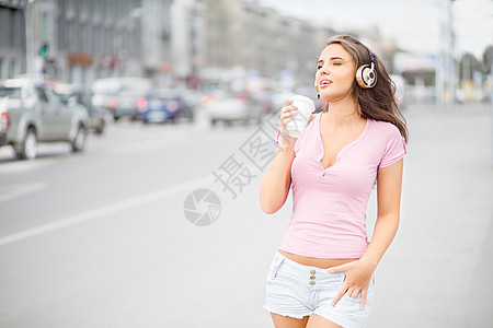 美丽的轻女人带着音乐耳机,着个走的咖啡杯,城市交通背景下闭着眼睛听音乐图片