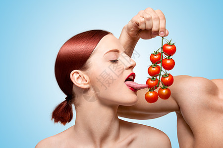 美丽的女孩用她诱人的舌头藤蔓上舔成熟的红色樱桃西红柿前戏与食物喂食蓝色背景图片