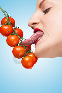美丽的女孩舔成熟的红色樱桃西红柿藤蔓上的感与她诱人的舌头蓝色的背景图片