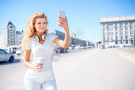 美丽的轻女人带着音乐耳机个带走咖啡杯,拍照自己,自拍城市背景图片