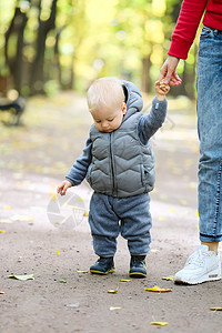 户外穿着温暖背心夹克的蹒跚学步的孩子的肖像岁的小男孩秋天的公园里学他的母亲散步图片