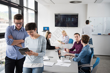 两个业务人员用平板电脑为下次会议准备,并与背景中的同事讨论想法图片