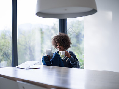 轻的非裔美国妇女微笑着坐明亮的窗户旁边,看着平板电脑,着白色的杯子她豪华的家图片
