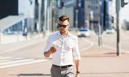 商业技术通信人的戴太阳镜的轻人带着智能手机城市里散步图片