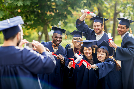 教育毕业技术人的群快乐的国际学生穿着灰浆板学士学位礼服,户外用智能手机拍照图片