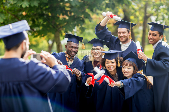 教育毕业技术人的群快乐的国际学生穿着灰浆板学士学位礼服,户外用智能手机拍照图片