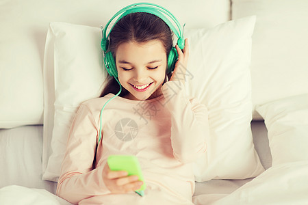 人们,孩子,休息技术快乐的微笑女孩躺床上,用智能手机耳机听家里的音乐快乐的女孩躺床上,家里智能手机图片