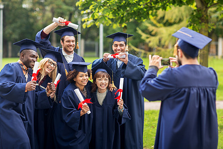教育毕业技术人的群快乐的国际学生穿着灰浆板学士学位礼服,户外用智能手机拍照学生单身汉用智能手机拍照图片