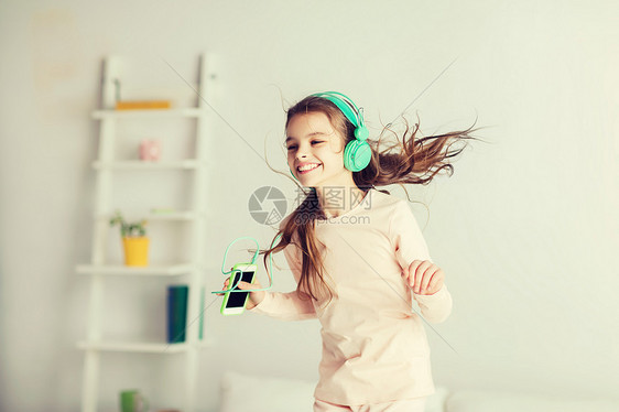 人们,孩子,睡衣派技术快乐的微笑女孩戴着耳机,用智能手机床上跳跃,家听音乐女孩用智能手机耳机床上跳跃图片