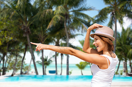 暑假,旅行,人度假的快乐的轻女人戴着帽子,指着异国情调的热带海滩,棕榈树游泳池的背景夏天海滩上戴帽子的快乐轻女图片