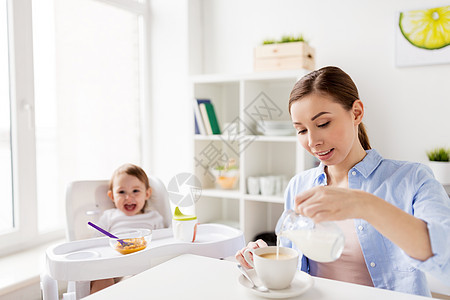 家庭,食物,饮食,早餐人们的快乐的母亲把牛奶水壶倒咖啡杯,小婴儿坐家里厨房的高椅子上快乐的母亲婴儿家里吃早图片