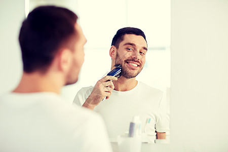 美容,卫生,剃须,美容人的轻人看着镜子剃须胡须与修剪机电动剃须刀家庭浴室男人浴室里刮胡子,修边器图片