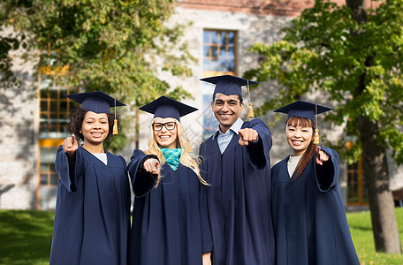 教育毕业人的群快乐的国际学生穿着迫击炮板学士学位服校园建筑背景上指着你快乐的学生单身汉指着你图片