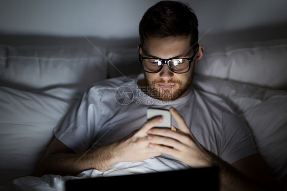 技术互联网通信人的轻人戴着眼镜,晚上家里卧室的床上着笔记本电脑智能手机晚上床上笔记本电脑智能手机的男图片