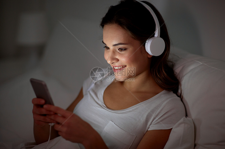 技术,休闲人的快乐的微笑轻妇女与智能手机耳机听音乐床上晚上女人床上智能手机耳机图片