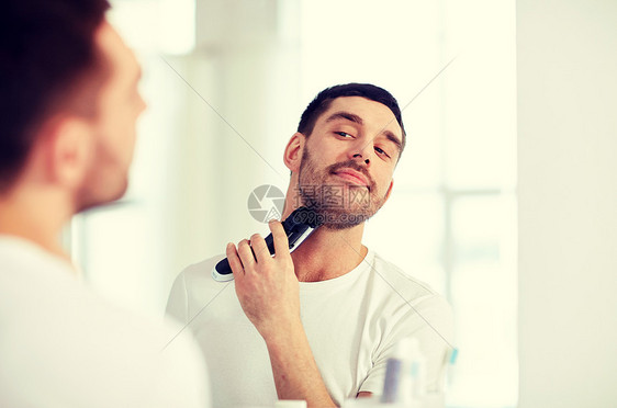 美容,卫生,剃须,美容人的轻人看着镜子剃须胡须与修剪机电动剃须刀家庭浴室男人浴室里刮胡子,修边器男人图片