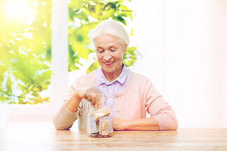 保险,退休人的微笑的老年女子把钱放家里的璃罐上,窗户上绿色的自然背景老妇女家把钱放进璃瓶里老妇女图片