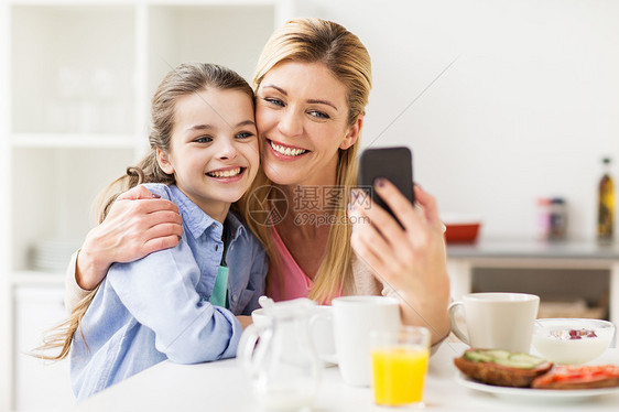 家庭,技术人的快乐的母亲女儿与智能手机吃早餐自拍家里的厨房幸福的家庭家里用智能手机自拍幸福的家庭家里用图片