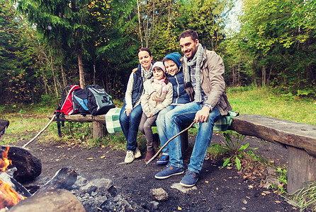 野营,旅游,徒步旅行人们的快乐的家庭坐长凳上的营地火灾树林里幸福的家庭坐篝火旁的长凳上图片