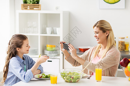 家庭人的快乐的母亲与智能手机吃饭拍照她的女儿家里的厨房女人家用智能手机拍摄女儿图片