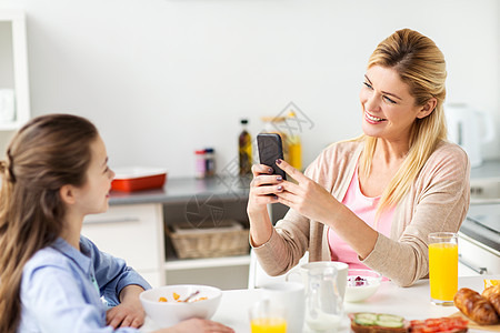 家庭,技术人的快乐的母亲与智能手机吃早餐拍照她的女儿家里的厨房女人家用智能手机拍摄女儿图片