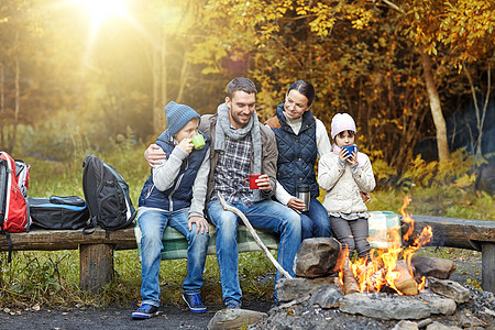 野营,旅游,旅游人们的快乐的家庭坐长凳上,喝着热茶杯子营地火灾秋天的森林幸福的家庭坐篝火旁的长凳上图片