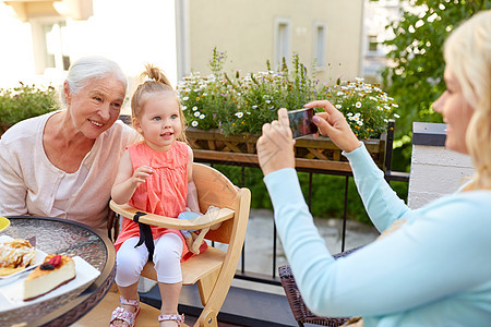 家庭世代人的快乐的微笑母亲与智能手机拍照女儿祖母咖啡馆餐厅露台女人咖啡馆给她的家人拍照图片