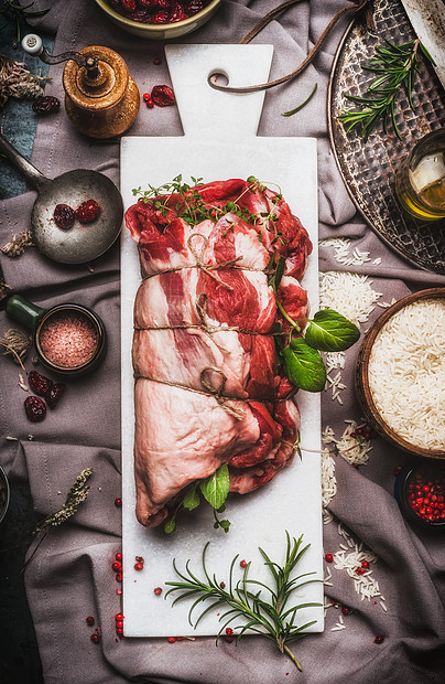 生羊肉肩烤与绳子,充满草药香料白色大理石切割板准备乡村背景与餐巾,米饭烹饪勺子,顶部视图肉类食品图片