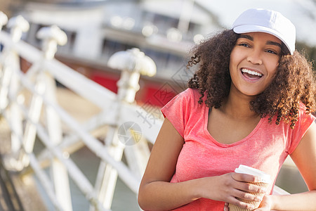 美丽快乐的混合种族非裔美国女孩,十几岁的女青,着完美的牙齿,微笑着喝着外卖咖啡,外戴着白色棒球帽粉红色的t恤图片