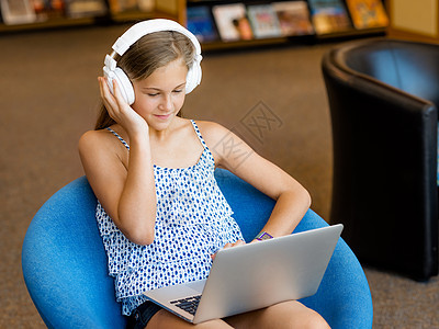 个带耳机的十几岁女孩坐图书馆里阅读倾听图片