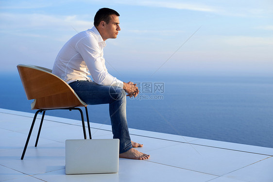 英俊的轻人放松工作笔记本电脑家里的阳台,同时看日落图片