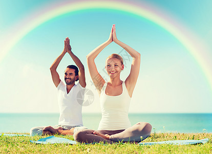 运动,健身,瑜伽人的微笑的夫妇冥想坐垫子上,举手海上彩虹的蓝天背景图片
