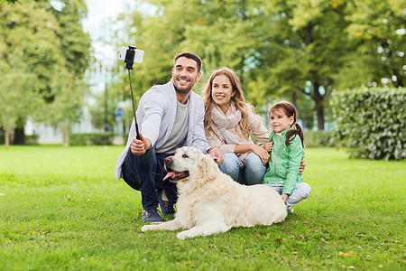 家庭,宠物,动物,技术人的快乐的家庭与拉布拉多猎犬狗公园的自拍杆上用智能手机拍照图片