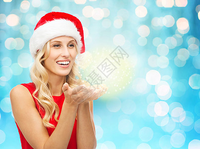 人们,假期,诞节魔法快乐的金发女人戴着诞老人的帽子,蓝色的灯光背景下,手掌上着仙女的灰尘图片
