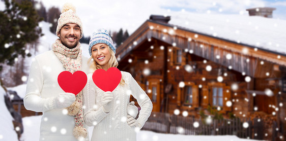 爱情,情人节,情侣,诞节人的微笑的男人女人戴着冬天的帽子围巾,木屋雪背景上着红色的纸心形图片
