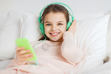 人们,孩子,休息技术快乐的微笑女孩躺床上,用智能手机耳机听家里的音乐图片