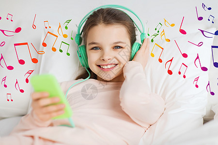 人,孩子技术快乐的微笑女孩躺床上,着智能手机耳机,家里听音乐,音符图片
