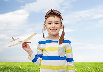 童,时尚人的快乐的微笑小男孩戴着飞行员的帽子,蓝天绿色的田野背景上玩木飞机图片