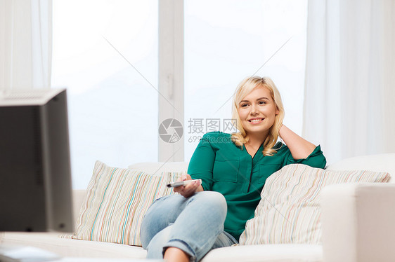 电视休闲人的微笑的女人坐沙发上,带着遥控器,家看电视图片