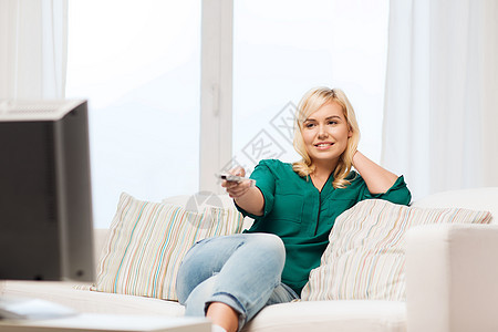 电视休闲人的微笑的女人坐沙发上,带着遥控器,家看电视图片