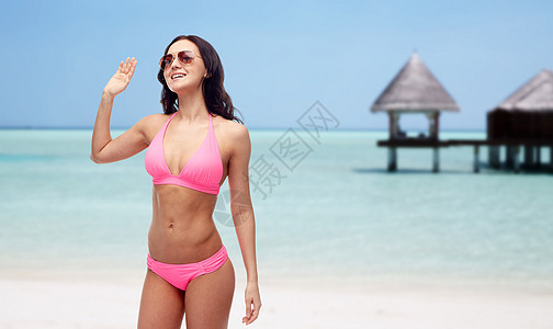 人们,泳装,夏季旅行手势快乐的轻女子戴着太阳镜粉红色泳衣,挥舞着手马尔代夫海滩上的平房背景图片