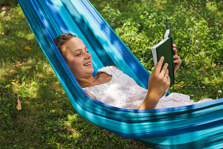 快乐的轻女孩蓝色的吊床上读本迷人的书,放松绿色的花园里用本书放松图片