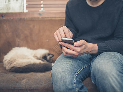 个轻人坐张旧沙发上,只猫他旁边睡觉,他正用他的智能手机图片