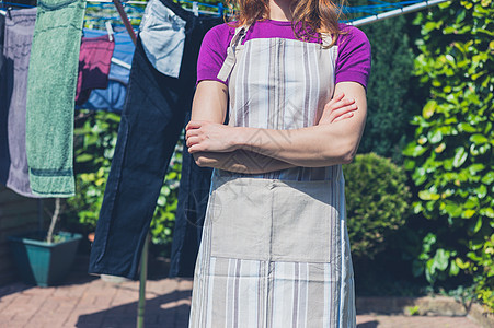 个穿着围裙的轻女人站她的洗衣房旁边,花园里的条晾衣绳上晾晒衣服图片