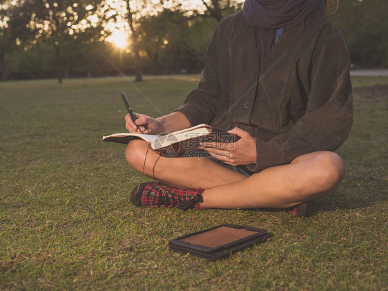 个轻的女人日落时坐公园的草地上,笔记本上写字,用平板电脑图片