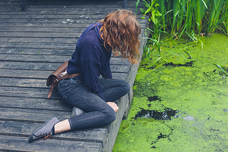 个轻的女人坐森林里藻类的肮脏池塘边的木甲板上图片