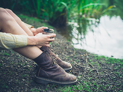 个轻的女人坐森林里的池塘边,正喝杯茶图片